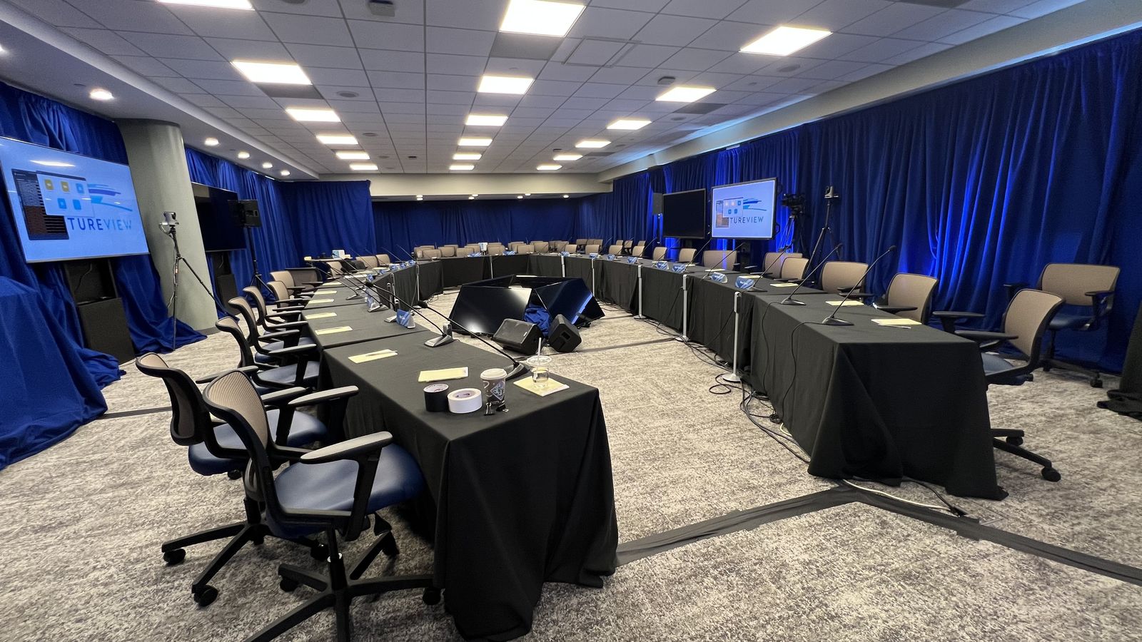 A hollow-square board room with microphones, displays and robotic cameras, framed in blue drape