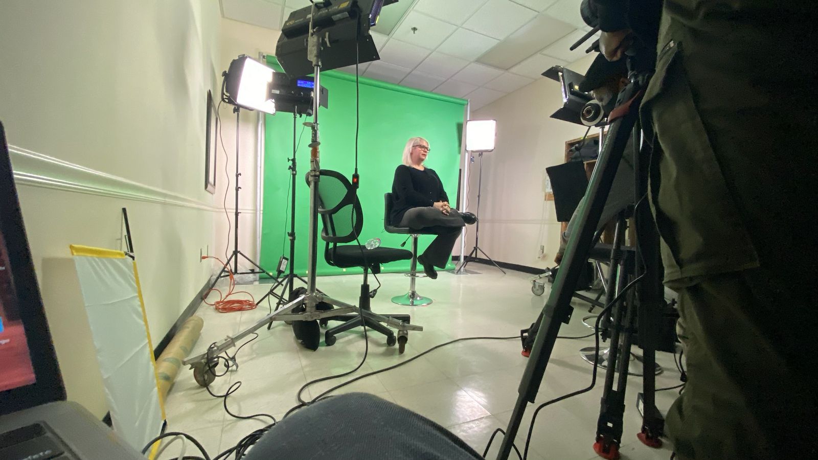 A presenter seated in front of a green screen, lit by LED panels, with a camera in the foreground