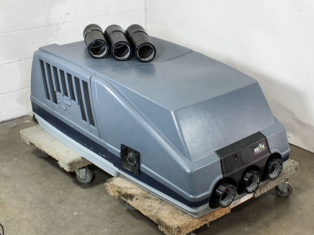 A vintage three-lens video projector on a wheeled dolly in a warehouse